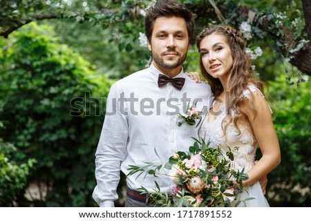 
Happy stylish couple the bride and groom are smiling and looking at the camera in the garden on their wedding day. The bride has a wedding bouquet in her hands. The groom wears a bow tie, the bride h Imagine de stoc © 
