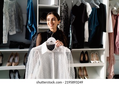 Feliz y elegante mujer mirando lejos y sonriendo mientras estaba de pie en el vestidor cerca del armario con muchos estantes y vistiéndose en casa