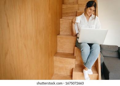 Happy stylish girl sitting on wooden stairs with laptop and holding cup of coffee. Young woman smiling , holding laptop, browsing, shopping online or working online from home. Stay home. Freelance - Powered by Shutterstock