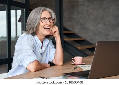 Happy Stylish Mature Middle Aged Business Woman Wearing Glasses Laughing Sitting At Workplace Desk With Laptop. Cheerful Positive Older Grey-haired Businesswoman Having Fun Working Alone In Office.