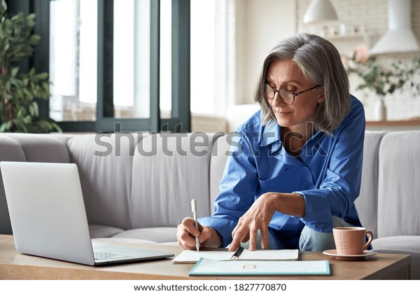 Happy stylish mature old woman remote working\
from home distance office on laptop taking notes. Smiling 60s\
middle aged business lady using computer watching webinar sit on\
couch writing in\
notebook.