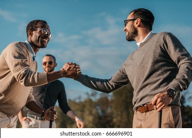 happy stylish multicultural friends shaking hands while playing golf on golf course 