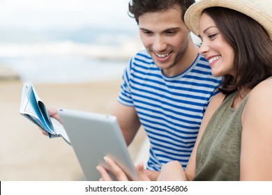 Happy Stylish Young Couple On Summer Vacation At The Beach Looking Up Directions On A Tablet Computer Smiling As They Compare A Map Held By The Husband As They Plan Their Sightseeing