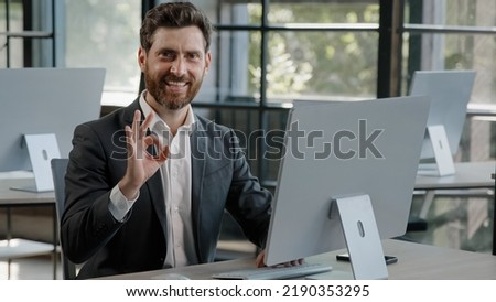 Happy success cheerful bearded 40s Caucasian adult man 30s businessman manager worker working with computer in office smile looking into camera with sign ok good idea done job approve agree