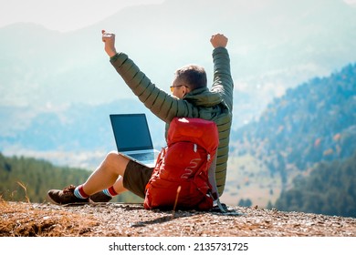 Happy Success Man Using Laptop And Raised Arms On Mountain Peak Celebrate His Freedom. Work Online. Man Works On Laptop. Tourist With Backpack.