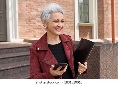 Happy Successful 50 Year Old Female In Trendy Jacket Posing On Doorway Holding Smart Phone And Folder, Having Cheerful Smile. Positive Mature Businesswoman Using Mobile Outdoors, Messaging Online