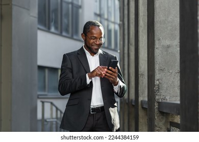 Happy successful African American businessman boss walking outside office building, manager holding smartphone reading message and smiling, using communication app.