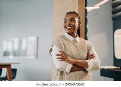 Happy and successful African businesswoman in a corporate office. She stands with crossed arms, deep in thought, looking away. Her pensive expression suggests she is reflecting on her work.