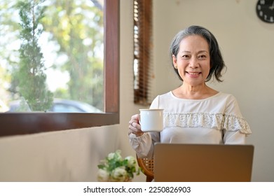 Happy and successful aged Asian businesswoman remote working at the cafe, sipping coffee while using laptop computer. smiling and looking at the camera. - Powered by Shutterstock