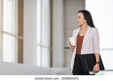 A happy successful Asian businesswoman holding a takeaway coffee cup, document file and digital tablet in the business building