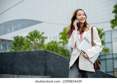 Happy successful Asian businesswoman holding laptop and Talk with cellphone on the Street next to a glass building. successfully work go to against city scene in background of modern office buildings. - Powered by Shutterstock