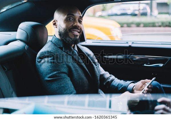 Happy successful bearded and bald black businessman\
in formal clothes looking at camera and using cellphone while\
sitting in car