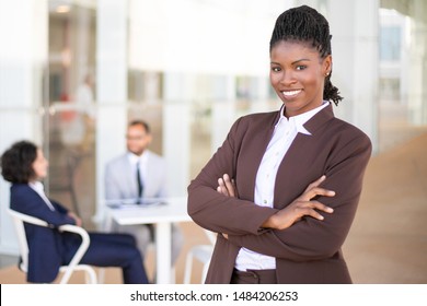 Happy Successful Business Leader Posing While Her Team In Background. Young African American Business Woman Standing With Arms Folded And Smiling. Teamwork Concept