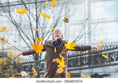 A happy and successful businessman in a brown coat throws yellow maple autumn leaves in a city park against the background of a business center, spreading his arms to the sides, made a successful deal
