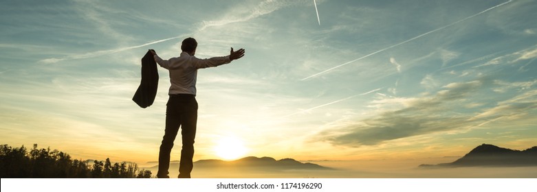 Happy successful businessman celebrating on a wall watching the sunrise silhouetted against the colorful orange sky in a panorama view with copy space.
