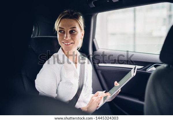Happy successful businesswoman with tablet for\
digital marketing looking away and smiling while sitting in luxury\
car and getting to destination with driver,positive female proud\
ceo holding touch pad