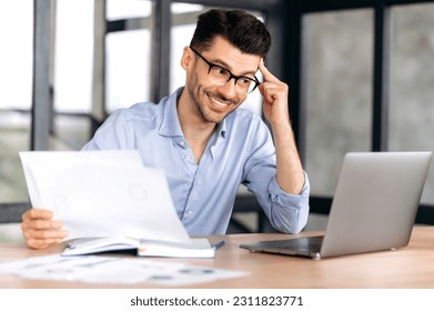 Happy successful confident young caucasian businessman, manager or freelancer wearing glasses sitting at table in office enjoying profit, holding business documents, looking at laptop screen, smiling - Shutterstock ID 2311823771