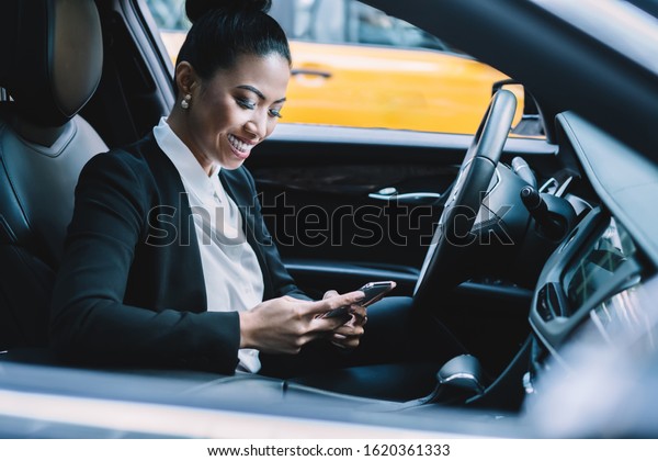 Happy successful executive charming Asian female\
manager in formal suit text messaging on cellphone while sitting\
during stop behind car\
wheel