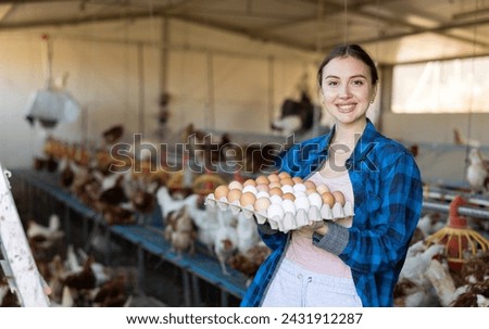 Happy successful female owner of poultry farm engaged in breeding of laying hens standing in chicken coop, holding carton tray of fresh organic eggs