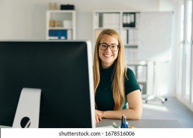 Happy successful friendly young business manageress seated at her desk looking around her monitor with a smile at the camera in a high key office