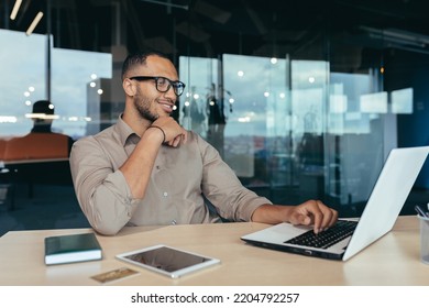 Happy and successful Indian man working in modern office building, programmer in glasses using laptop to write code, businessman in shirt smiling and happy