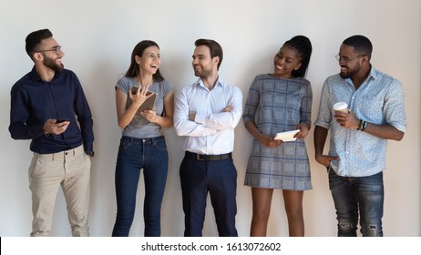 Happy successful multiethnic millennial employees stand in row talk laugh in office hall, smiling young diverse multiracial young people or colleagues gather near white wall show unity and team spirit
