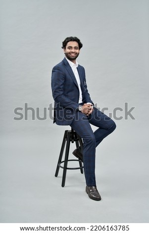 Happy successful rich young indian business man ceo leader, wealthy arab professional manager, confident male businessman executive wearing suit sitting on chair isolated on beige, vertical portrait.