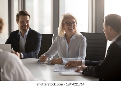 Happy Successful Well-dressed Company Members Headed By Aged Woman In Eyeglasses Gathered Together In Boardroom At Briefing. Business Partners Solve Current Matters Negotiating Sitting At Office Desk