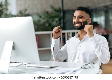 Happy Successful Young Adult Indian Business Leader, Manager Or Office Worker With Beard Sitting In Office, Using Computer, Rejoicing In Success Or Profit, Gesturing With Hands, Smiling