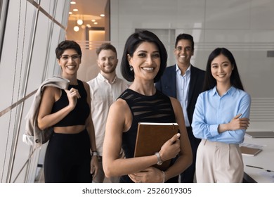 Happy successful young Middle Eastern project leader woman posing with for portrait, standing on front of diverse business team, looking at camera, enjoying leadership, group support - Powered by Shutterstock