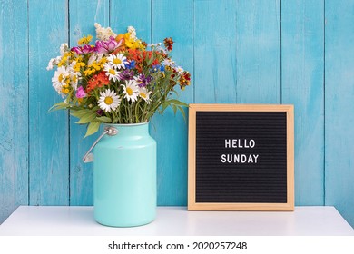 Happy Sunday words on black letter board and bouquet of bright wildflowers in tin can vase on table against blue wooden wall. Concept Hello Sunday.
