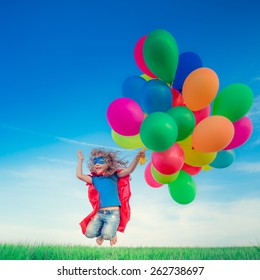 Happy Superhero Child Jumping With Colorful Toy Balloons Outdoors. Smiling Kid Having Fun In Green Spring Field Against Blue Sky Background. Freedom Concept