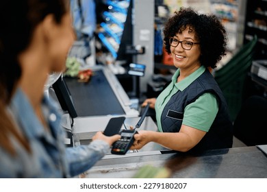 Happy supermarket cashier accepting mobile payment from a customer at store checkout.