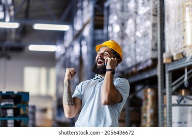 A happy supervisor is standing in the warehouse and talking on the phone with a boss. They sold a big amount of goods and he is cheering and holding a fist because of this success. - Powered by Shutterstock