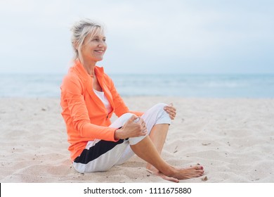 Happy supple mature blond woman seated in the golden sand on a beach looking to the side of the frame with a beaming smile against an ocean backdrop - Powered by Shutterstock