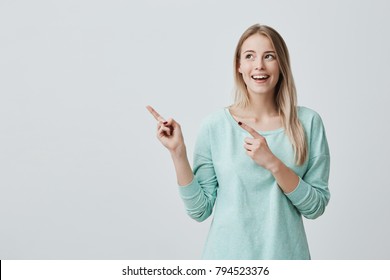 Happy surprised blonde young female smiling broadly at camera, pointing fingers away, showing something interesting and exciting on studio wall with copy space for your text or advertising content