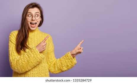 Happy Surprised Cute Woman Looks With Curious Gaze Points Aside At Free Space, Demonstrates Something At Upper Right Corner, Suggests Visit New Store, Invites To Get Inside, Wears Yellow Jumper