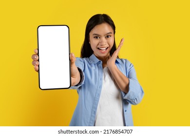 Happy Surprised Millennial Asian Woman Making Hand Gesture And Showing Smartphone With Empty Screen Isolated On Yellow Background, Studio. Win Online, Great Offer And Sale, New App, Tech And Website