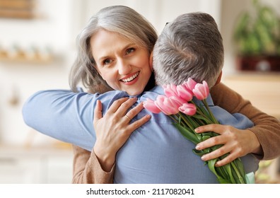 Happy Surprised Senior Woman Embracing Husband While Receiving Bouquet Of Fresh Spring Flowers Tulips On Womens Day At Home, Selective Focus. Elderly Married Couple In Love Celebrating Valentine's Day