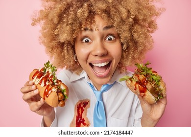 Happy Surprised Womsan Eats Delicious Hot Dog And Hamburger With Ketchup Prefers Fatty High Calorie Food Enjoys Taste Dressed In Formal Outfit Isolated Over Pink Background. Cheat Meal Concept