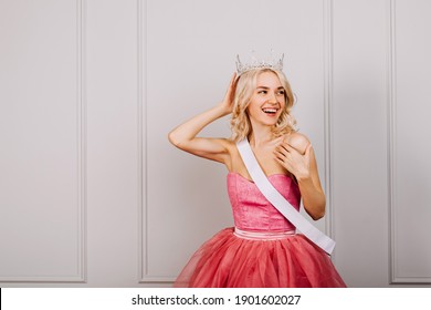 Happy Surprised Young Blonde Woman Wearing A Crown, Pink Dress And A White Ribbon, Smiling. Beauty Contest Winn