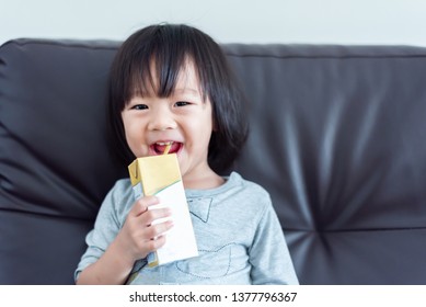 Happy Sweet Asian Baby Child Drinking A Carton Of Milk From Box With Straw On Sofa