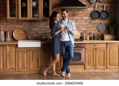 Happy sweet barefooted young couple drinking coffee in stylish kitchen together, holding mugs, hugging, talking, laughing, enjoying morning leisure time and hot beverage at home. Full length