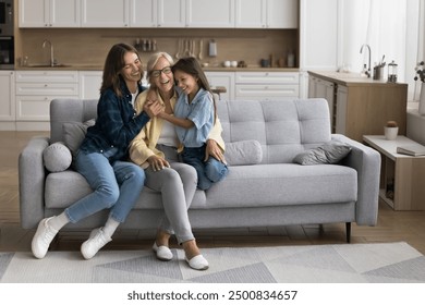 Happy sweet kid girl and young mom hugging cheerful grandma with love, support, laughing, smiling, sitting on sofa at home, enjoying family meeting. Three female generations shot - Powered by Shutterstock