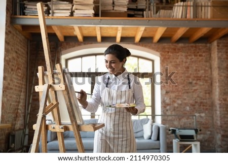  Happy talented young Indian artist girl drawing, painting on canvas with brush, pallet, working on artistic grunge retro designed home studio interior, feeling inspired, excited, smiling
