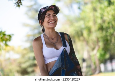 Happy and tattooed girl with cap, denim dungarees and white crop top smiling and laughing in the park in a sunny day