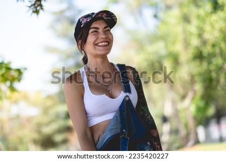 Happy and tattooed girl with cap, denim dungarees and white crop top smiling and laughing in the park in a sunny day