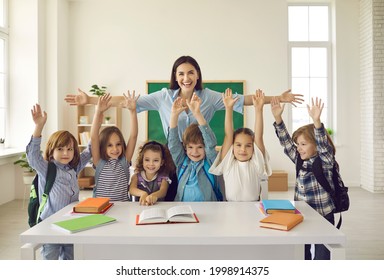 Happy teacher with arms widely spread to hug cheerful children raising hands. Positive portrait in classroom. Educator showing love, adoration and care for pupils. Children and tutor looking at camera