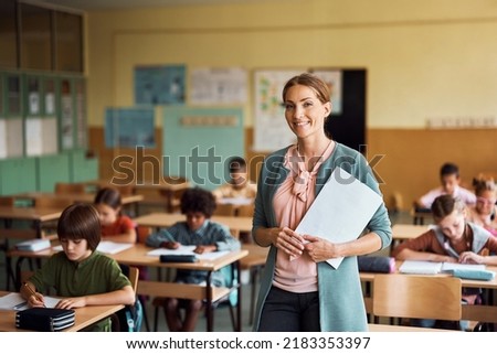 Happy teacher during a class at elementary school looking at camera. Her student are learning in the background.