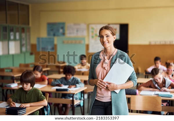 Happy teacher\
during a class at elementary school looking at camera. Her student\
are learning in the\
background.